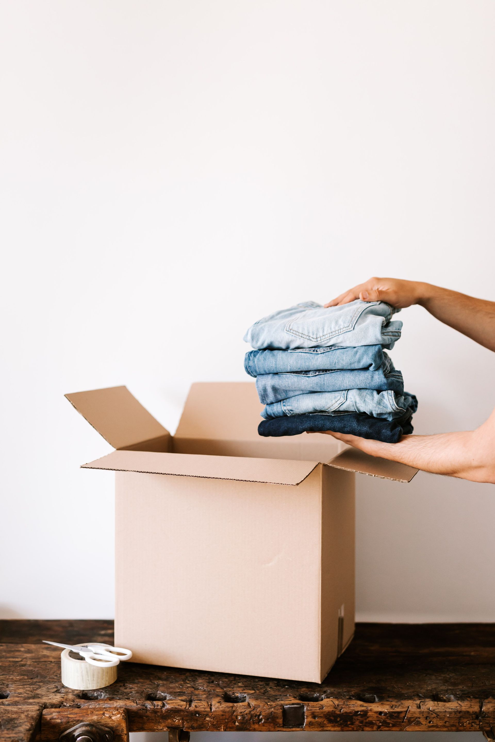 Crop unrecognizable person putting stacked jeans into cardboard box placed on wooden table against white wall before relocation